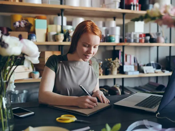 Female florist wearing apron writing in notebook beside laptop and fresh flowers in bright workshop for Town Service Net